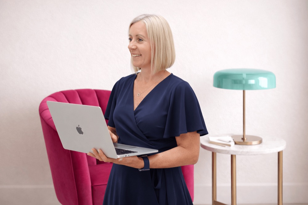 Laura with her laptop beside a raspberry chair and teal lamp.